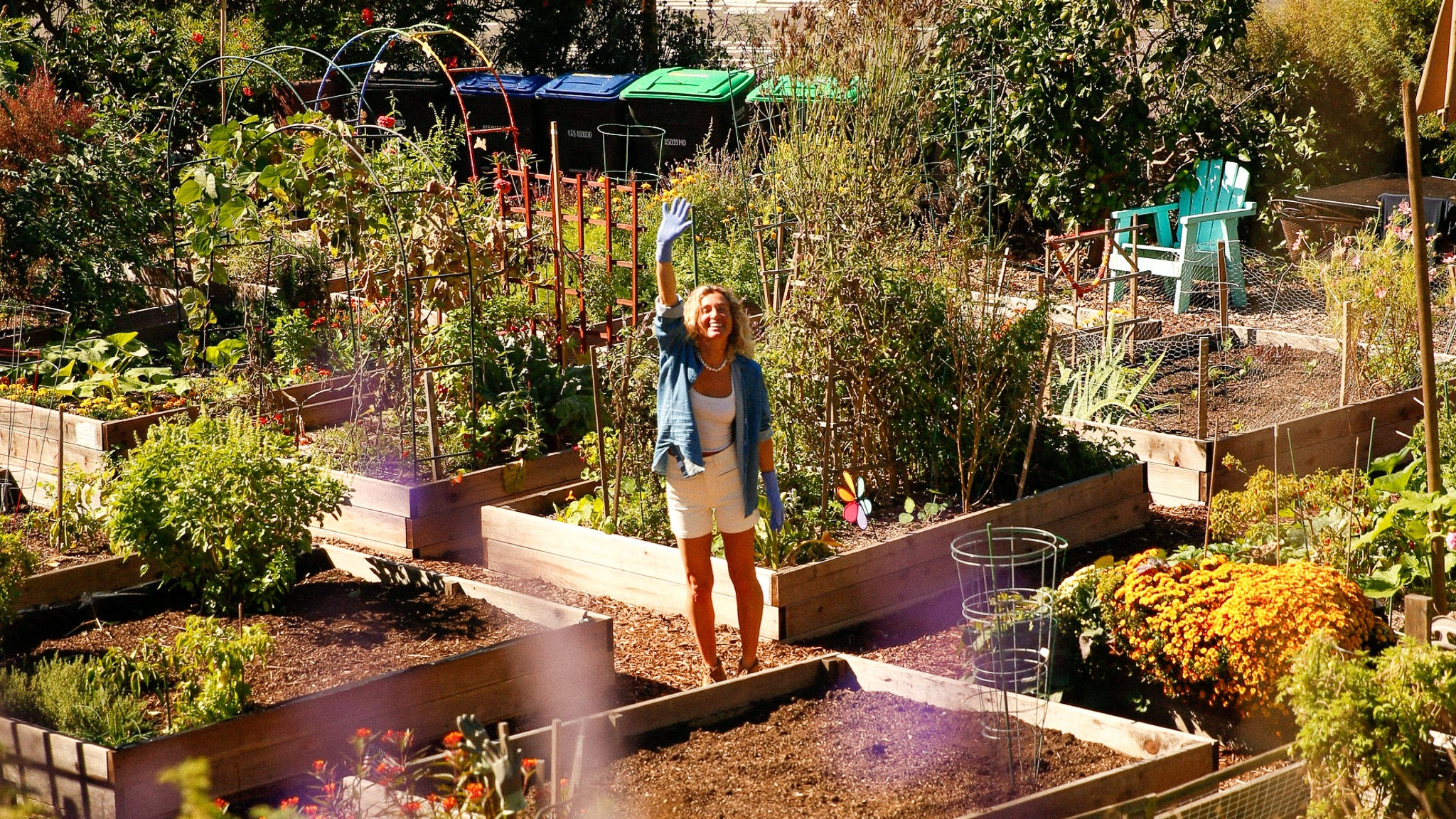 Woman standing in a community garden with raised beds and plants waving hi!