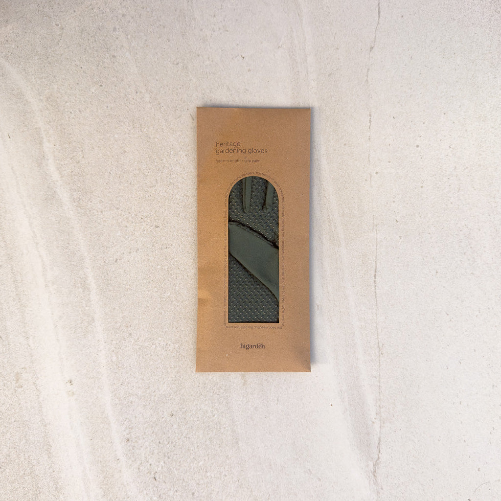 A rectangular kraft paper package with a cutout window displaying a dark olive green knit fabric item, photographed from above on a light textured surface.