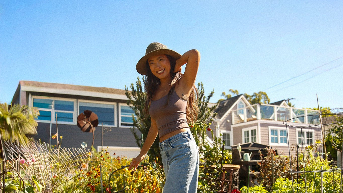 Woman wearing a handmade sun hat while gardening in a vibrant garden filled with flowers and greenery. Crocheted sunhat handmade in Bali.