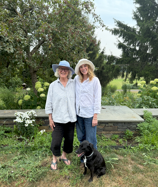 Two women and a dog standing in a garden with trees and flowers in the background