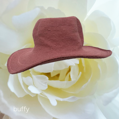 A small dusty rose knit hat with a wide brim, styled on top of a large white flower. The brand name 'buffy' is printed in white text in the bottom-left corner.