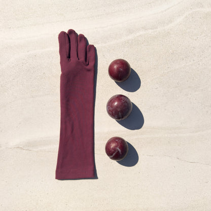 A single long stretch glove in deep burgundy is laid flat on a light beige sandy surface, next to three small round plum-like fruits with matching shadows.