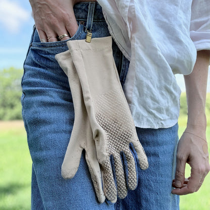 A person wearing blue jeans and a white shirt holds a pair of beige gardening gloves with textured grip palms against their waist, with a blurred outdoor background.