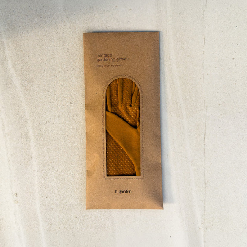 A single tan knit glove displayed inside a vertical kraft paper sleeve with a die-cut window, placed on a light textured surface. The sleeve has printed text including 'heritage gardening gloves' and the brand name 'hagardis'.