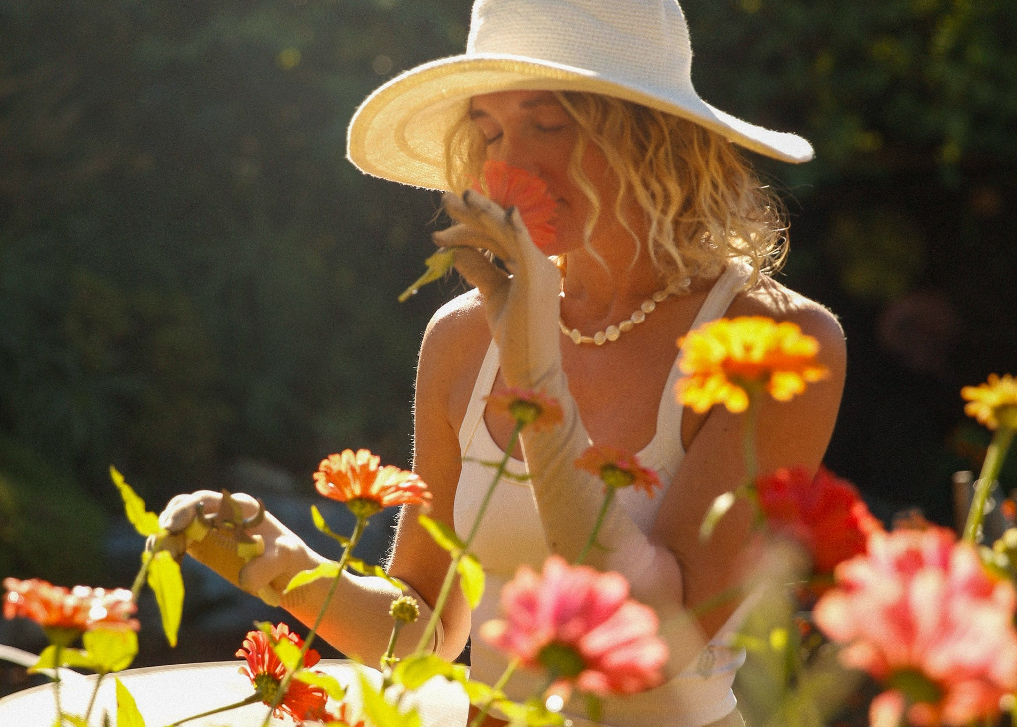 A woman wearing a bali wide-brim hat gently holds a flower while surrounded by colorful blossoms. Crocheted sunhat handmade in Bali.