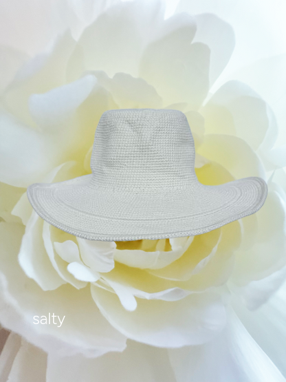 A light beige woven sun hat with a wide brim, displayed on top of a large white flower. The word 'salty' is printed in white text in the bottom-left corner.