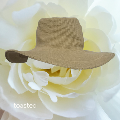 A small woven tan hat with a wide brim, resting on the petals of a large white flower. The word 'toasted' appears in white text in the lower-left corner.