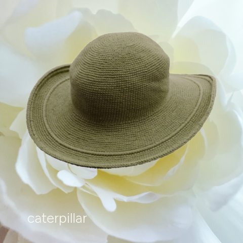 A small woven sun hat in olive green, displayed on a large white flower. The word 'caterpillar' is printed in white text in the bottom-left corner.