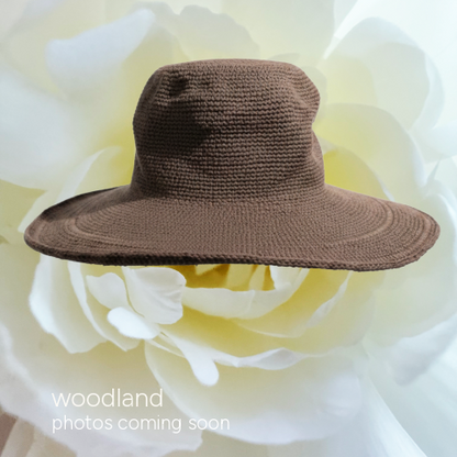A brown woven sun hat with a wide brim, displayed against a large white flower backdrop. Text in the lower-left corner reads 'woodland photos coming soon'.