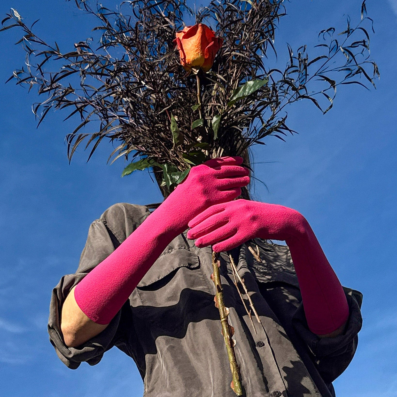 Person wearing pink gloves holding a bundle of sticks with a red rose against a blue sky.