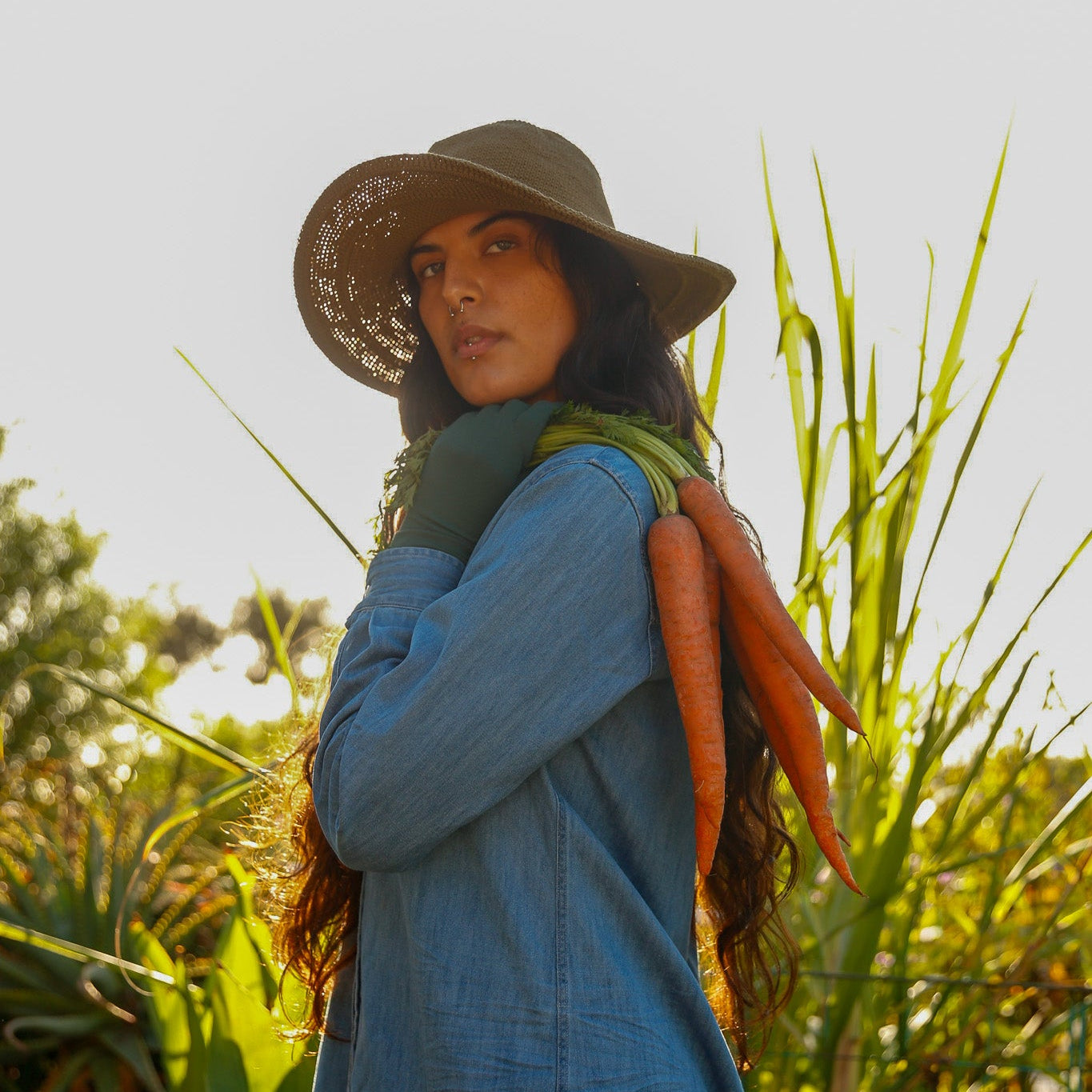 Woman holding carrots in a field wearing a wide-brimmed hat and blue dress.